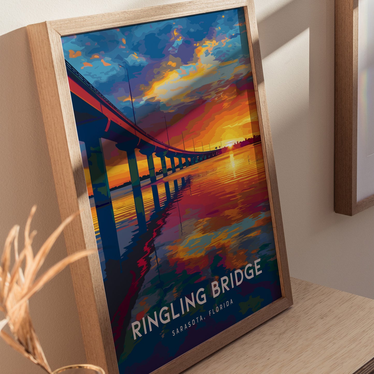 A framed poster depicting the John Ringling Bridge in Sarasota, Florida is displayed on a wooden surface. The artwork features a vibrant sunset with reflections on the water, capturing the bridges sweeping arc against a colorful sky.