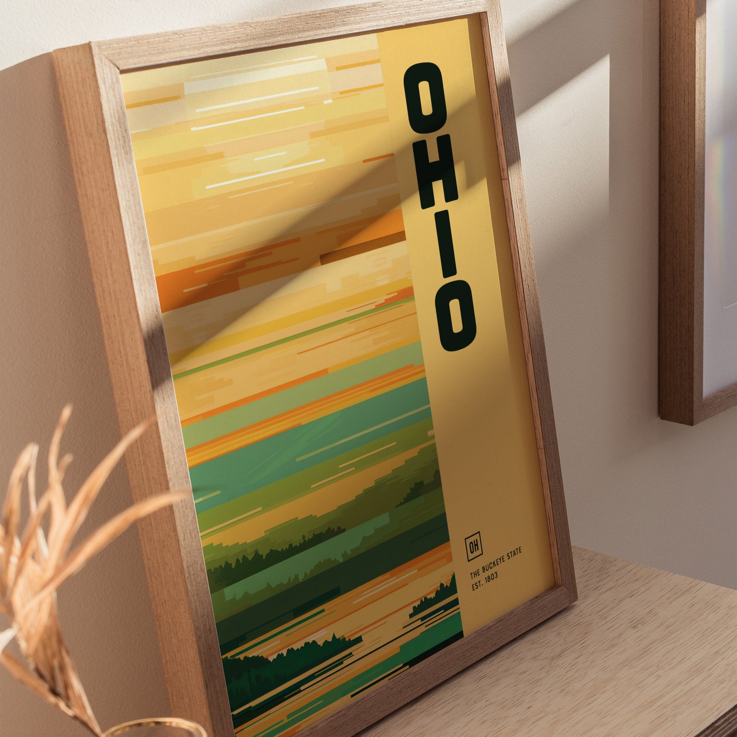 A framed Ohio poster with a minimalist design features bold vertical text and abstract landscape colors, displayed on a wooden surface with soft natural lighting.