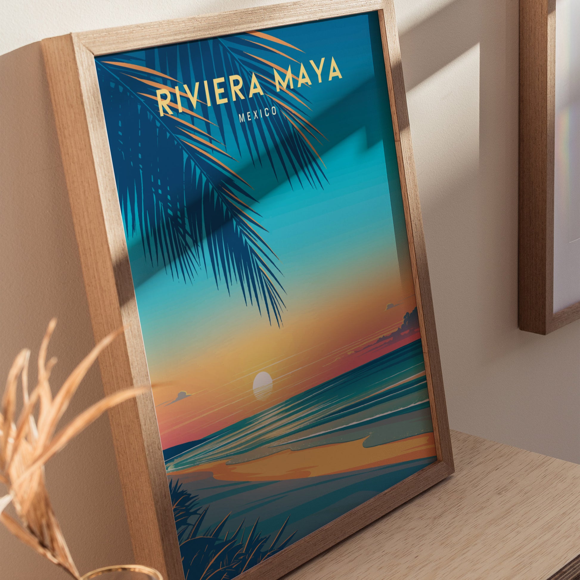 A framed poster of the Riviera Maya features a vibrant sunset over a beach with palm leaves in the foreground. The artwork is displayed on a wooden surface.