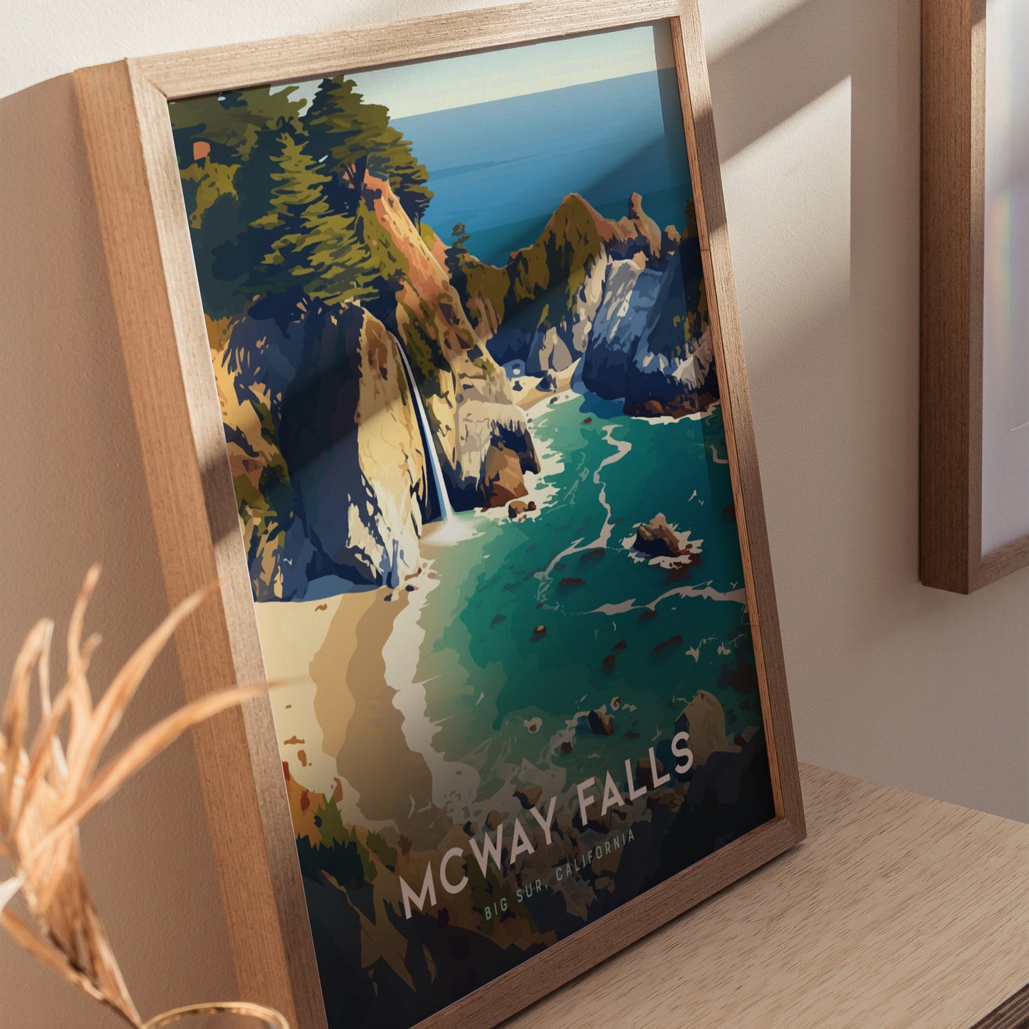 A framed poster of McWay Falls in Big Sur, California, features vibrant turquoise waters, rugged cliffs, and lush greenery, displayed on a wooden surface with soft lighting.