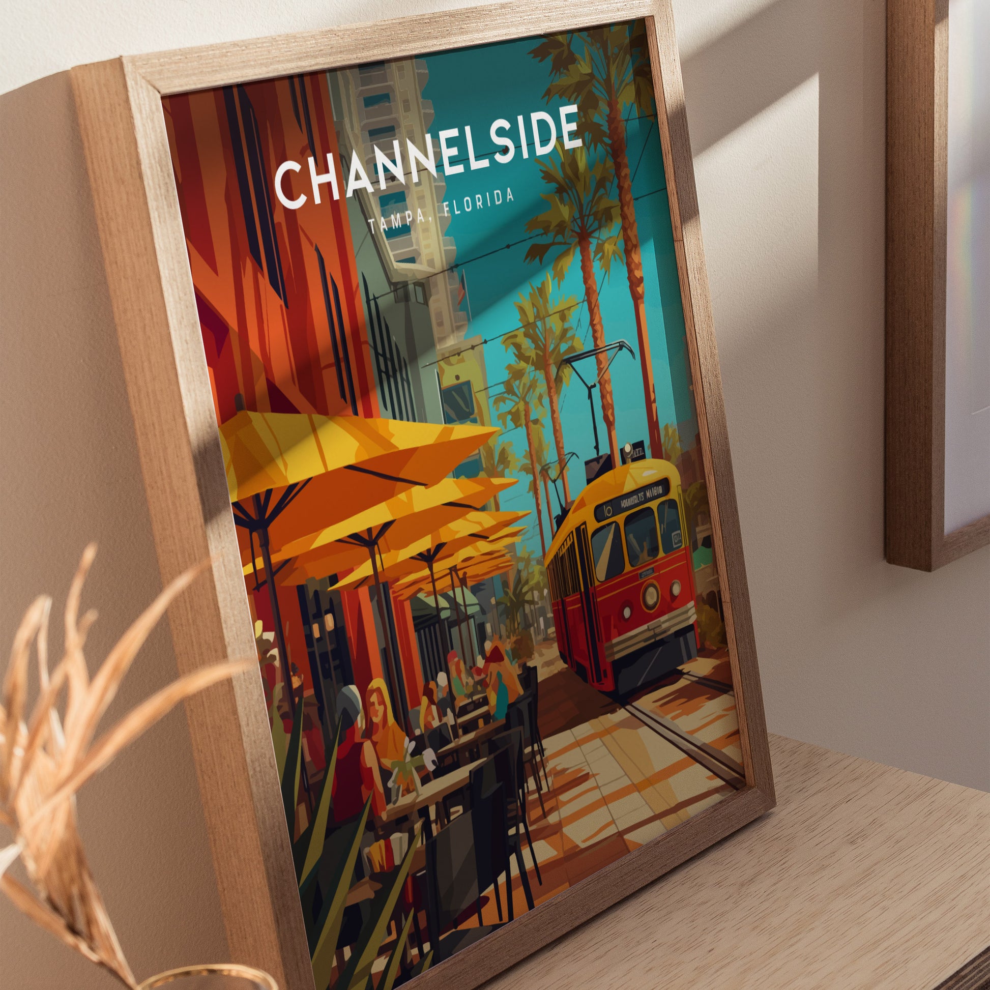 Framed Channelside Tampa poster featuring a lively street scene with a red trolley, yellow umbrellas, and people dining outdoors under palm trees. Bright, colorful depiction of Tampa, Florida.