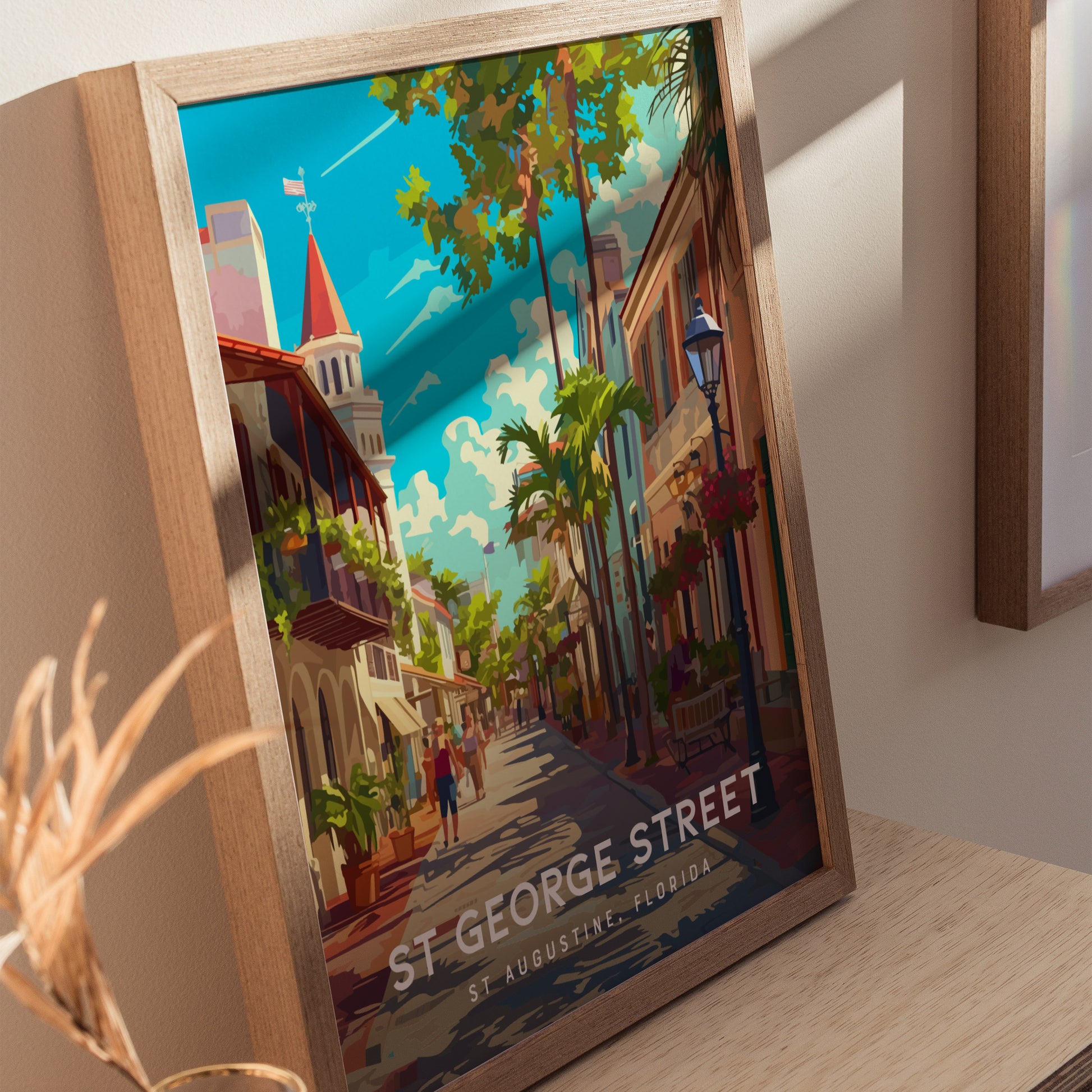 A framed poster depicts a vibrant, colorful scene of St. George Street with historic buildings, lush trees, and people strolling. The artwork is set against a bright, blue sky.