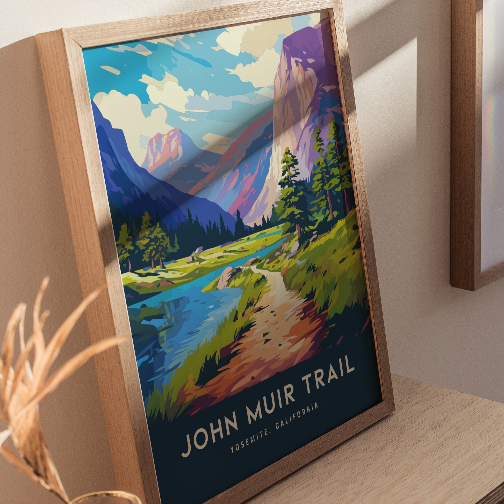 Framed John Muir Trail poster featuring vibrant illustrated landscape with mountains, trees, and a winding path. The artwork is propped against a wall on a wooden surface.
