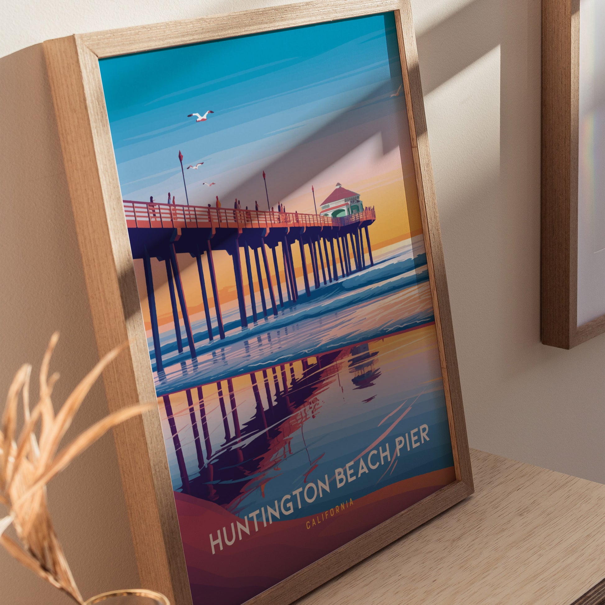 Framed poster of Huntington Beach Pier at sunset, featuring a long wooden pier with a pavilion, vibrant sky, and ocean reflections. Seagulls fly above, enhancing the serene scene.