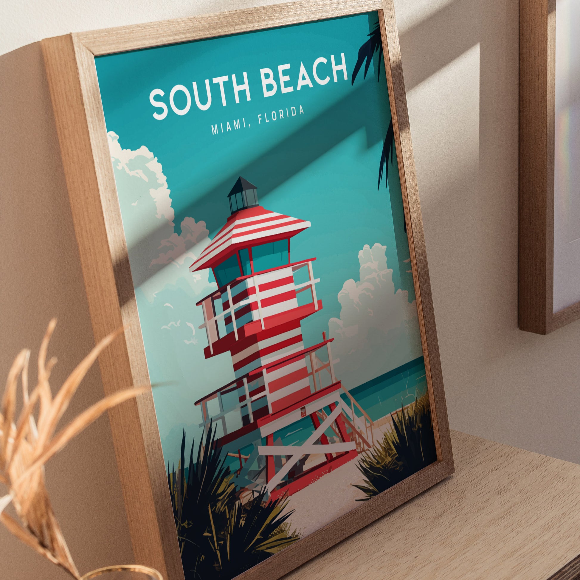 The image shows a framed poster of a South Beach lifeguard tower with bold red and white stripes, set against a turquoise sky and ocean. The artwork is displayed on a wooden surface, capturing the essence of Miami, Florida.