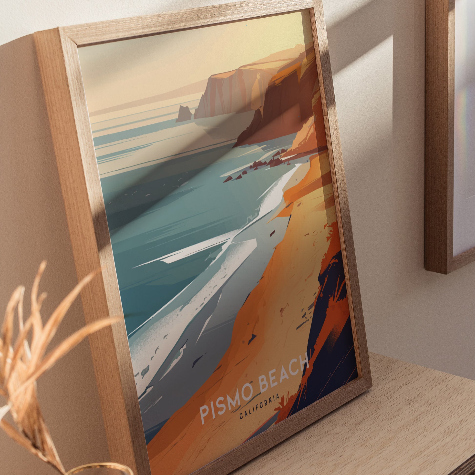 Framed artwork depicting a stylized Pismo Beach with vibrant orange cliffs, turquoise waters, and gentle waves under a sunset sky. The poster rests on a wooden surface.