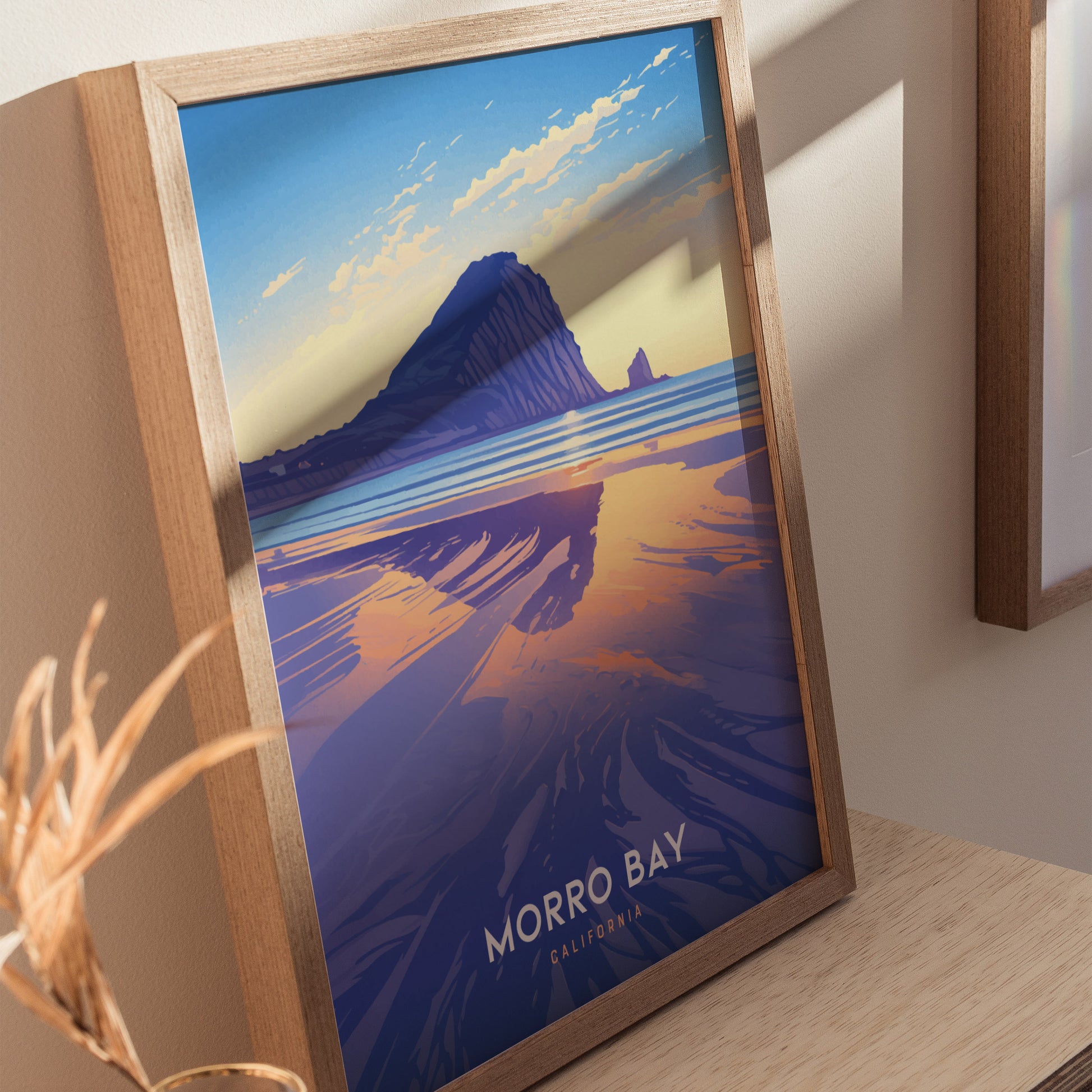 Framed artwork of Morro Bay, featuring Morro Rock with vibrant sunset colors reflecting on the water. The poster is propped on a wooden surface, casting a soft shadow.