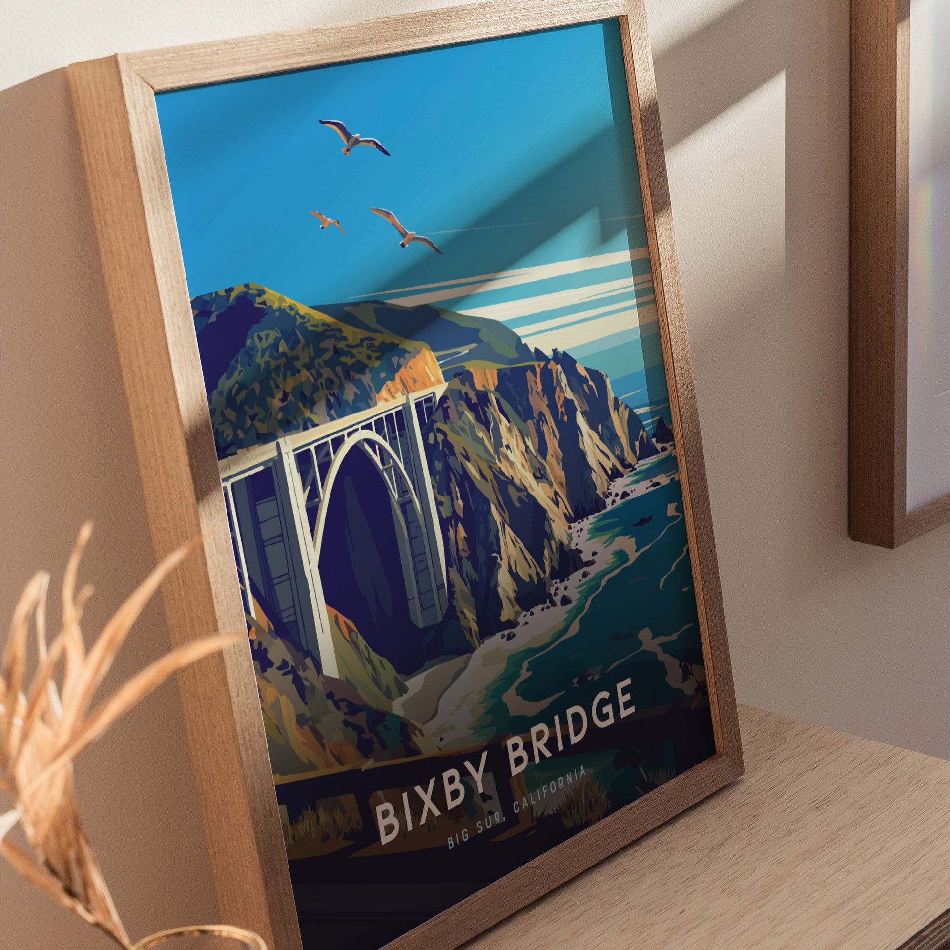 Framed poster depicting Bixby Bridge in Big Sur, California, showcasing the bridge spanning rugged cliffs with turquoise waters below and three birds flying in a clear blue sky.