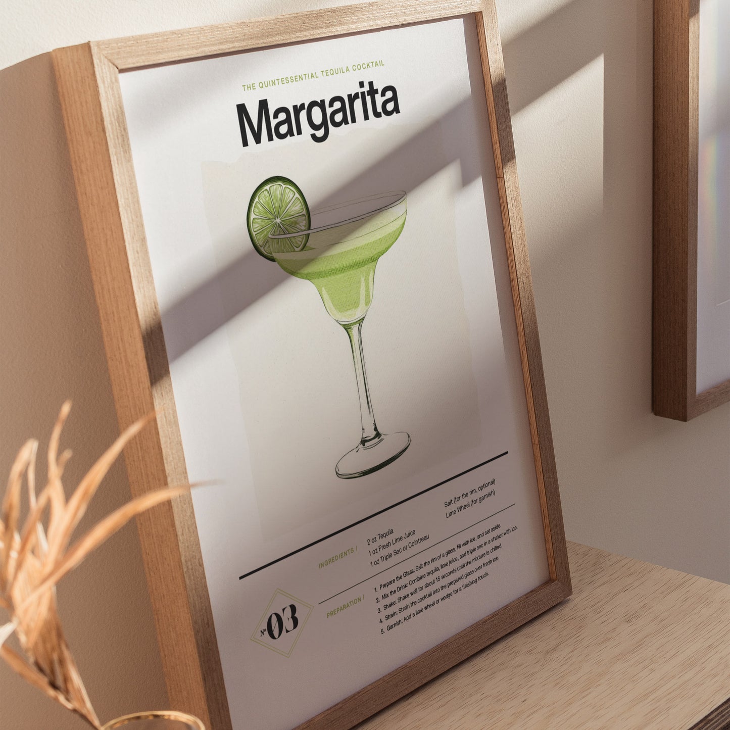 Framed Margarita cocktail poster featuring a detailed glass illustration with a lime slice, resting on a light wooden surface. Sunlight casts soft shadows on the wall.