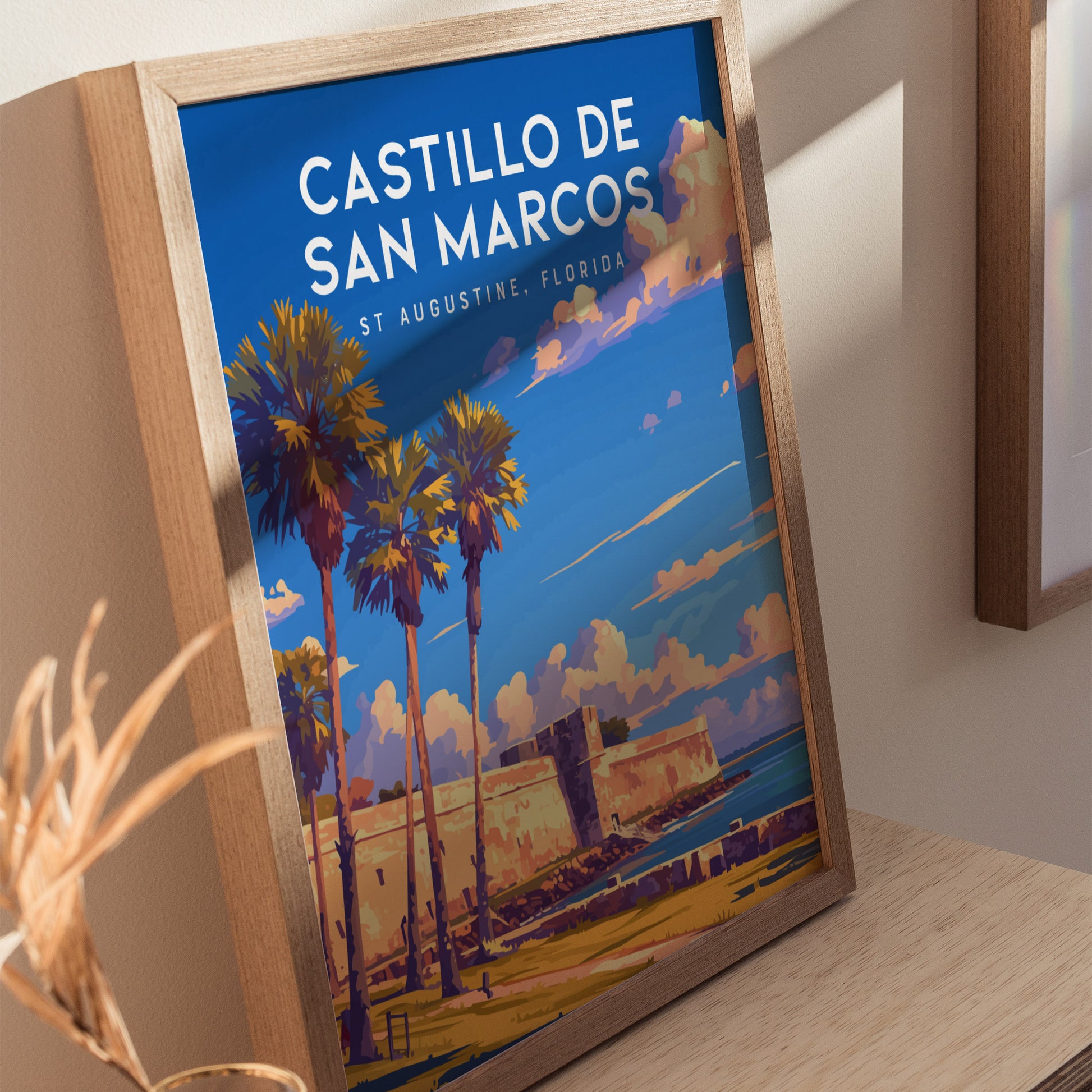 Framed poster of Castillo de San Marcos in St. Augustine, Florida, featuring palm trees and a vibrant sky, displayed on a wooden surface.