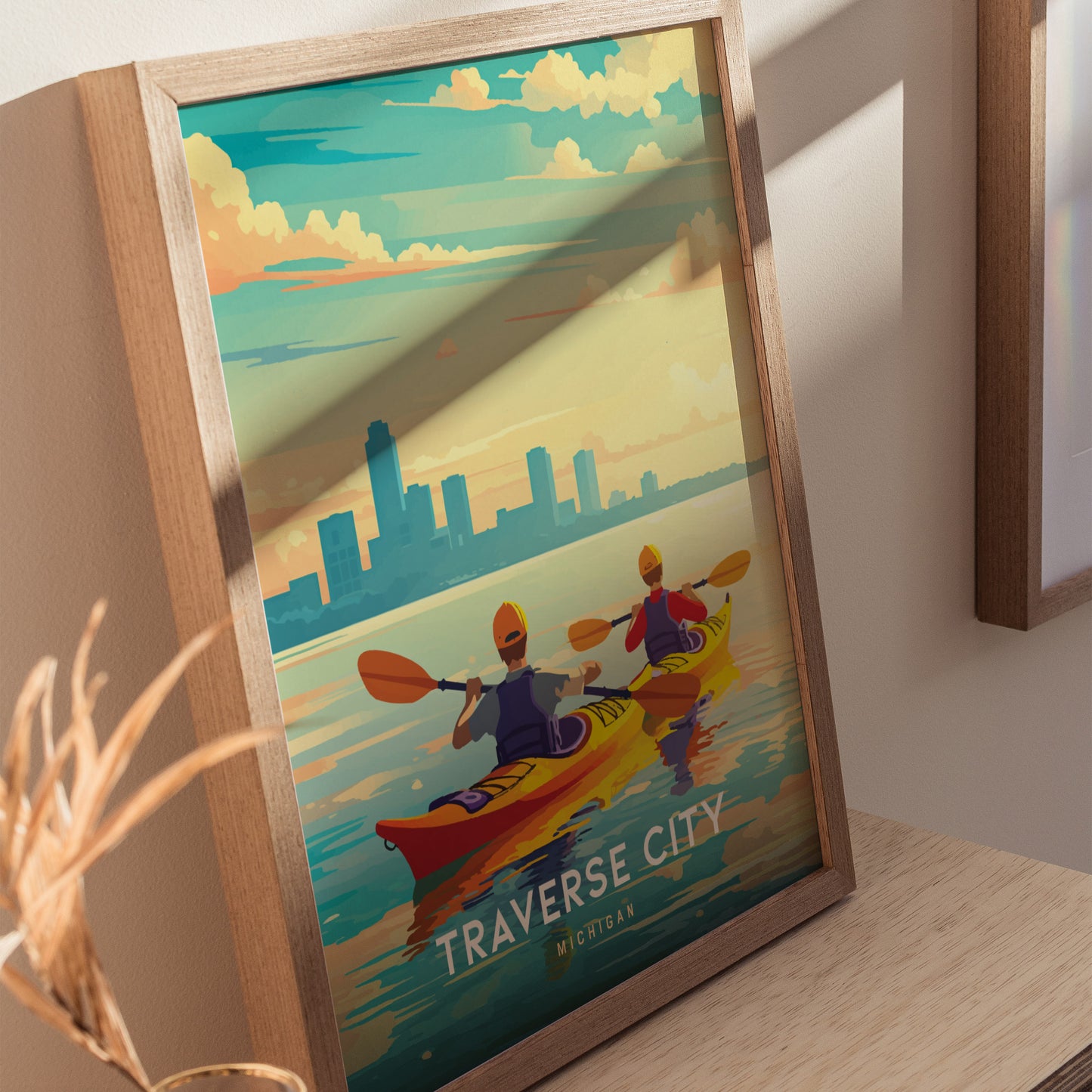 A framed Traverse City poster shows two kayakers paddling on serene water, with a city skyline and colorful sky in the background. The artwork captures a peaceful outdoor scene.