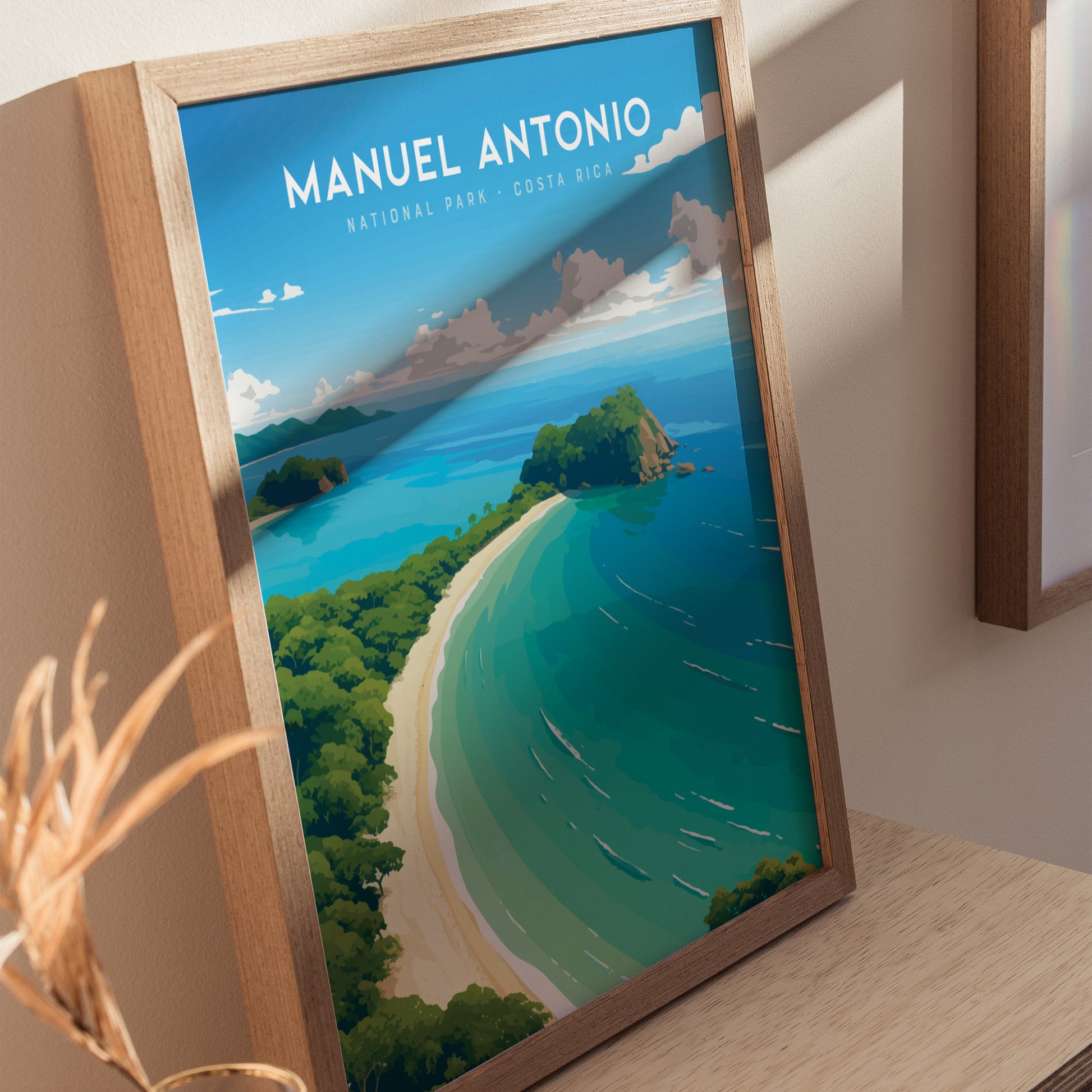 Framed poster of Manuel Antonio National Park featuring a vibrant illustration of a beach with turquoise water, lush green rainforest, and blue sky, displayed on a wooden table.