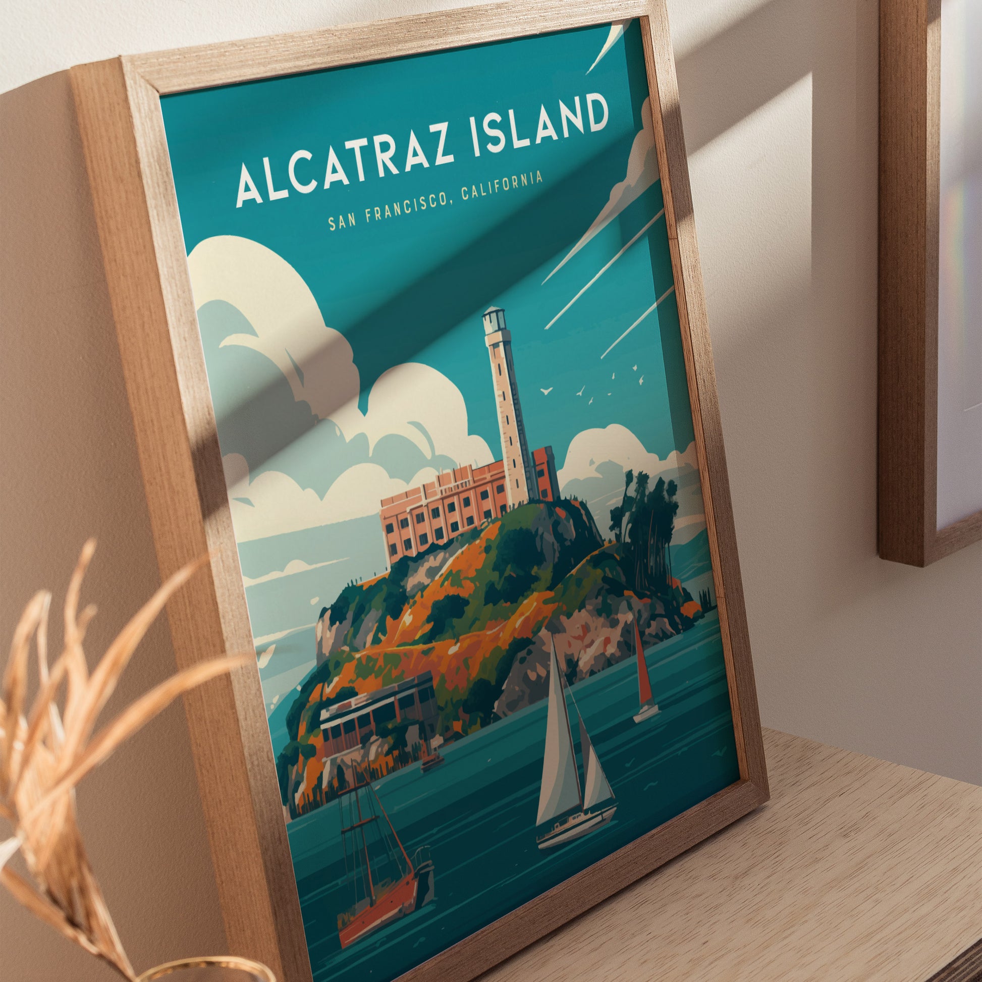 Framed poster depicting Alcatraz Island with a lighthouse, surrounded by ocean and sailboats under a blue sky with clouds. Displayed on a light wooden surface.