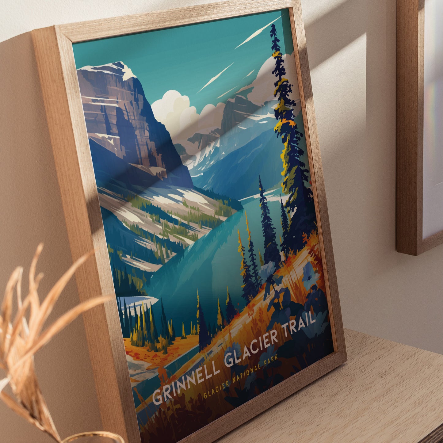 Framed artwork depicting the Grinnell Glacier Trail, featuring vibrant alpine meadows, turquoise lakes, and towering peaks under a clear sky. Positioned on a wooden surface, bathed in sunlight.