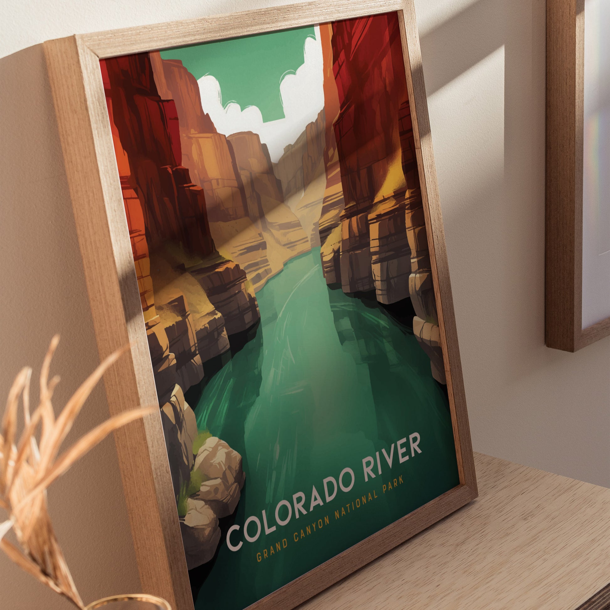 A framed poster depicting the Colorado River winding through the Grand Canyon with vibrant red and orange cliffs under a clear sky, placed on a wooden surface.