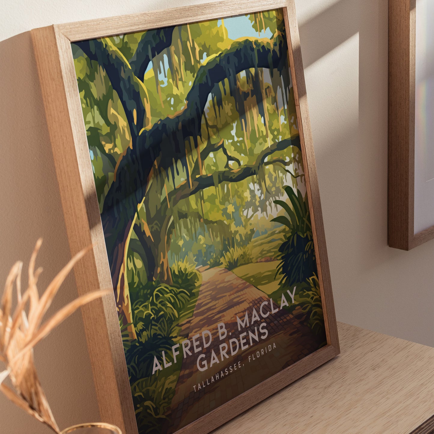 A framed poster of Alfred B. Maclay Gardens in Tallahassee, Florida, features vibrant moss-draped trees over a winding brick path, displayed on a wooden surface with soft lighting.