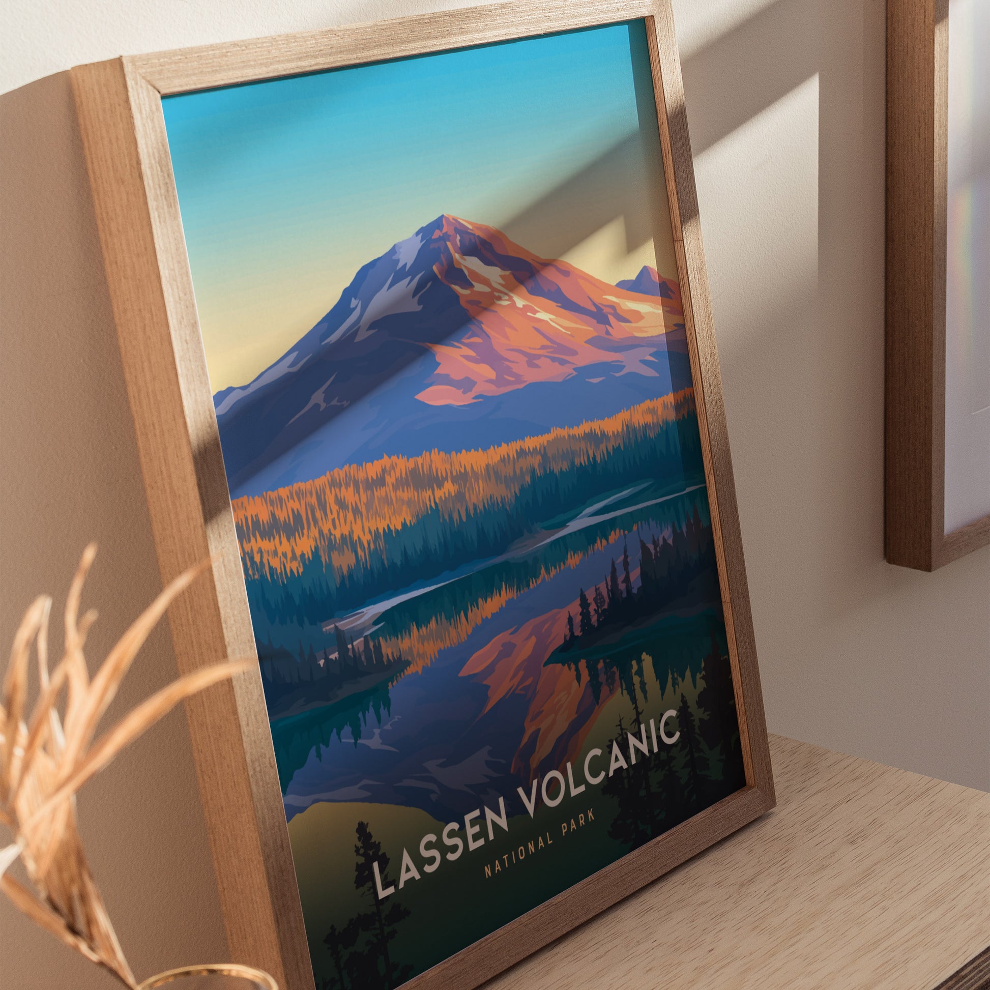 A framed poster of Lassen Volcanic National Park sits on a wooden surface, depicting a colorful landscape with a snow-capped mountain and vibrant forest reflected in water.