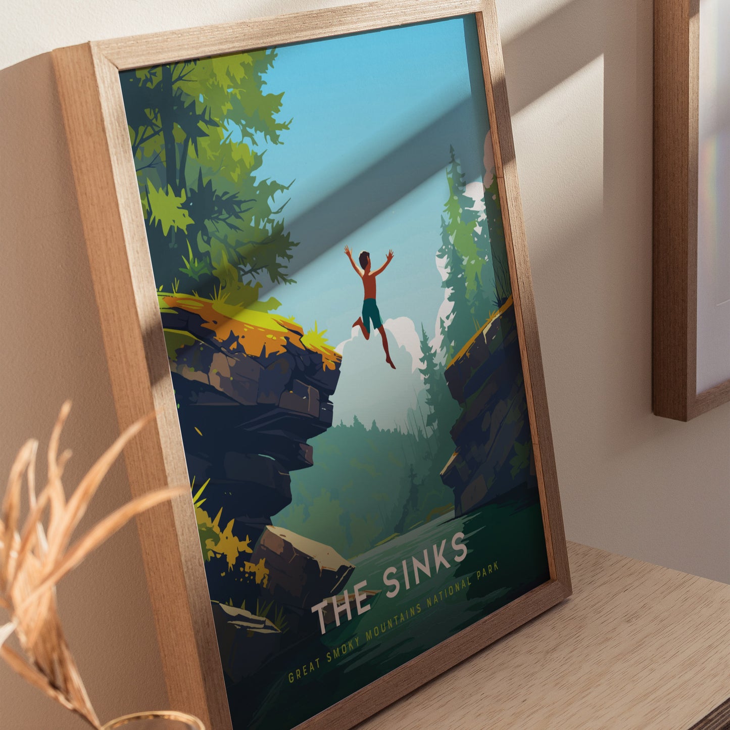 A framed poster titled The Sinks depicts a person joyfully leaping off a rocky ledge into a lush, forested canyon with a river below, set against a clear blue sky. The vibrant artwork captures the adventurous spirit of nature.