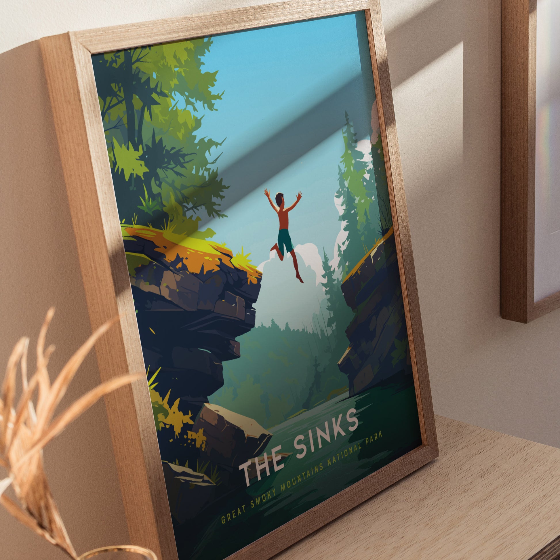 A framed poster titled The Sinks depicts a person joyfully leaping off a rocky ledge into a lush, forested canyon with a river below, set against a clear blue sky. The vibrant artwork captures the adventurous spirit of nature.