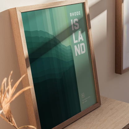 Framed abstract wall art featuring layered green tones with the text RHODE ISLAND on the right. The artwork is displayed on a wooden surface with soft lighting.
