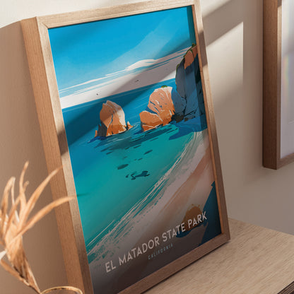 Framed poster of El Matador State Park featuring vibrant blue ocean, rocky cliffs, and clear skies, displayed on a wooden surface with soft natural lighting.
