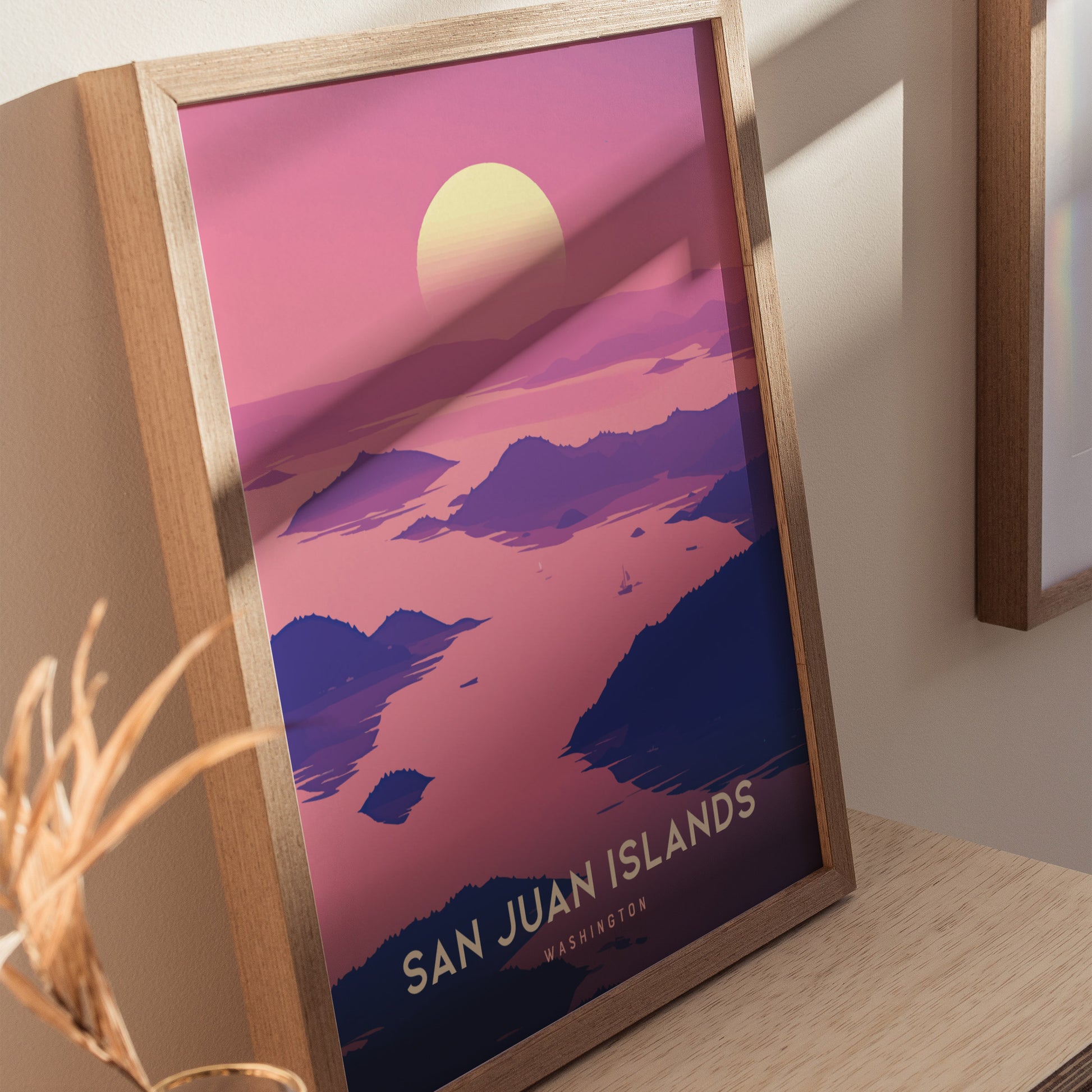 A framed poster of the San Juan Islands features a sunset over pink and purple hues, depicting serene island silhouettes. The artwork rests on a wooden surface.