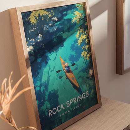 Framed artwork featuring an illustration of a kayaker on turquoise waters, surrounded by lush greenery. The poster is titled Rock Springs, Apopka, Florida and rests on a wooden surface.
