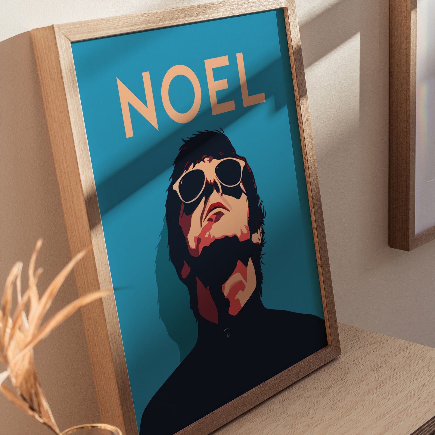 A framed poster features a stylized portrait of a person wearing sunglasses, with NOEL in bold letters above. The artwork rests on a wooden surface.