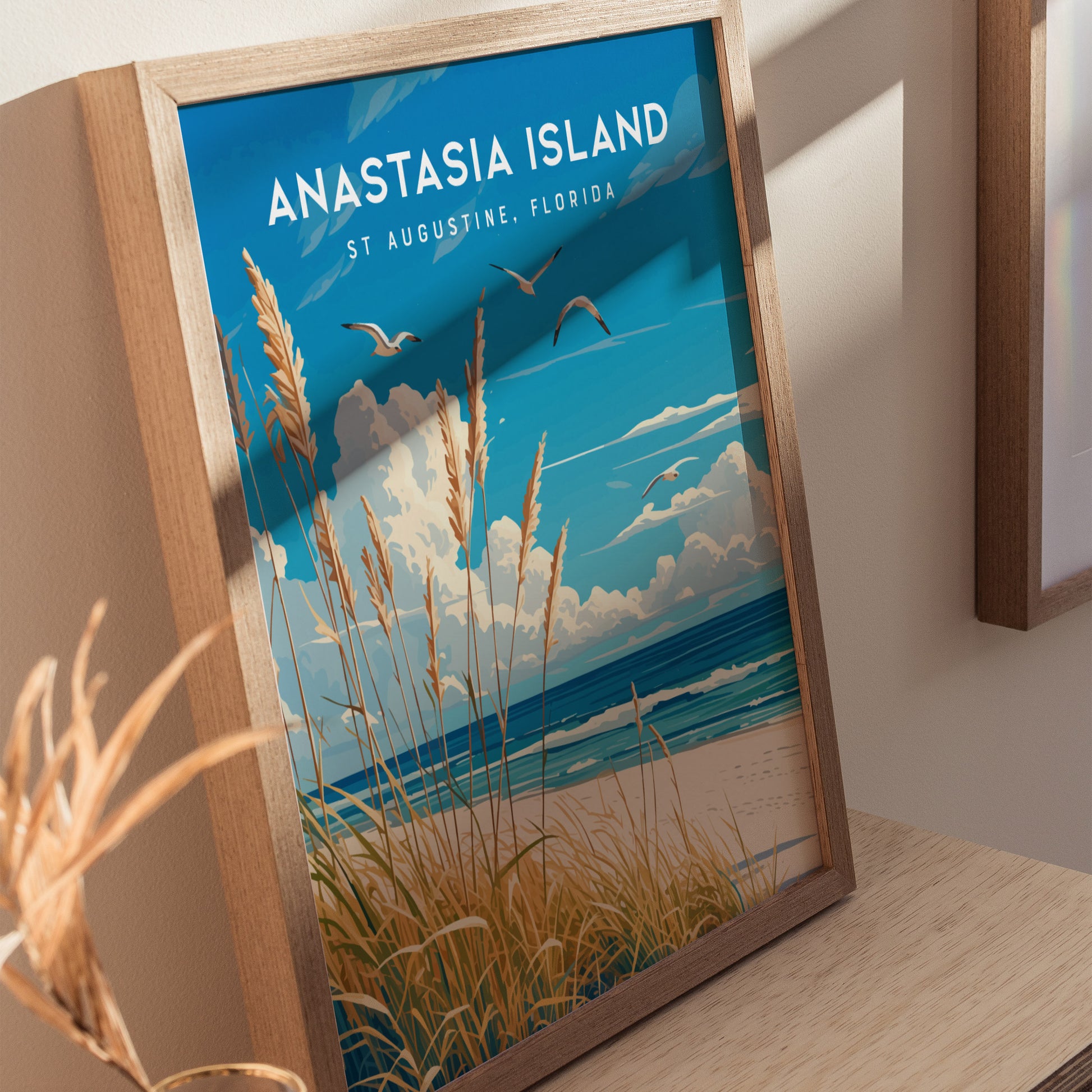 A framed poster of Anastasia Island in St. Augustine, Florida, featuring sea oats, a blue sky, and seabirds over the ocean. Its displayed on a wooden surface.