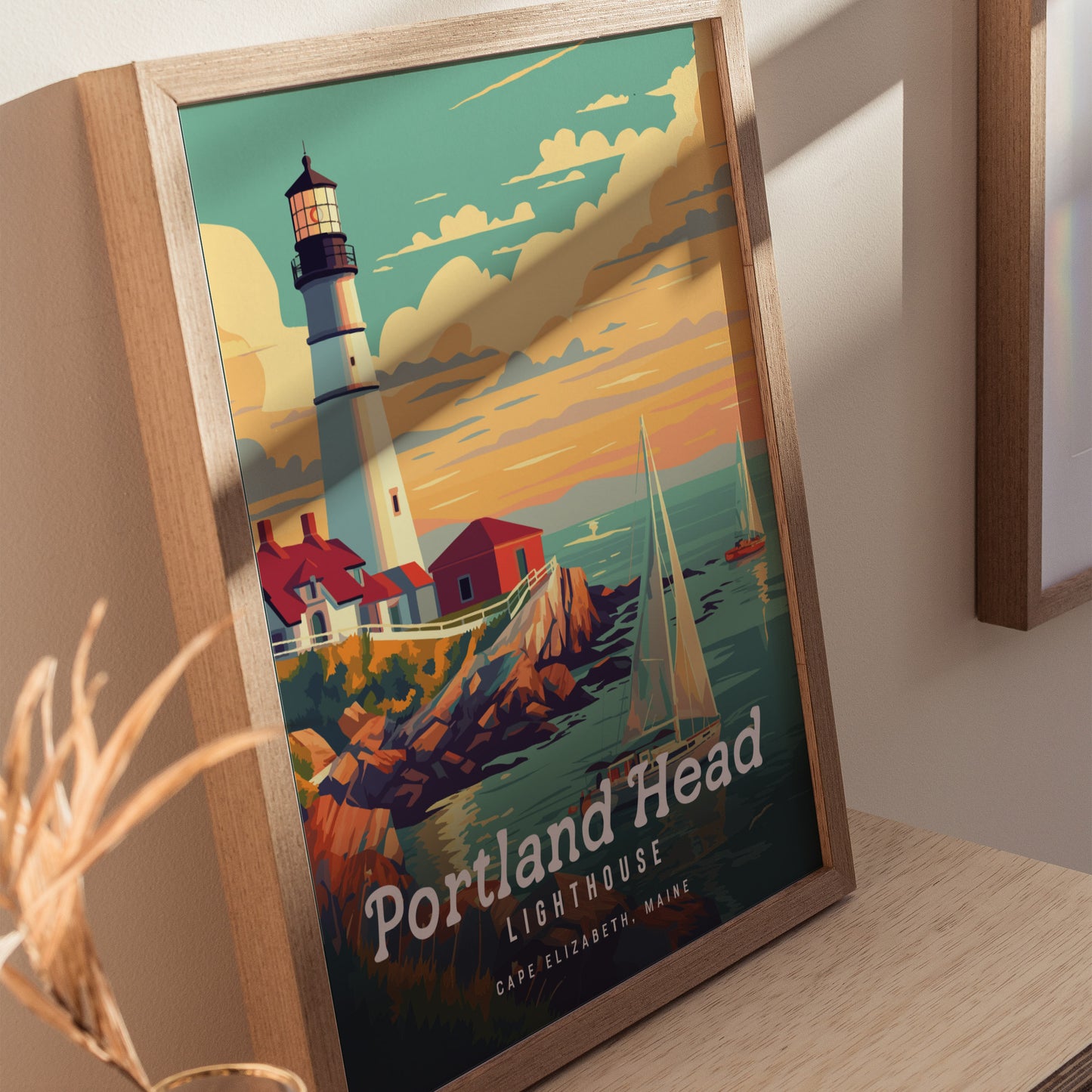 Framed poster depicting Portland Head Lighthouse with a white tower and red-roofed buildings on rocky cliffs. A sailboat glides on the ocean under a vibrant sunset sky.