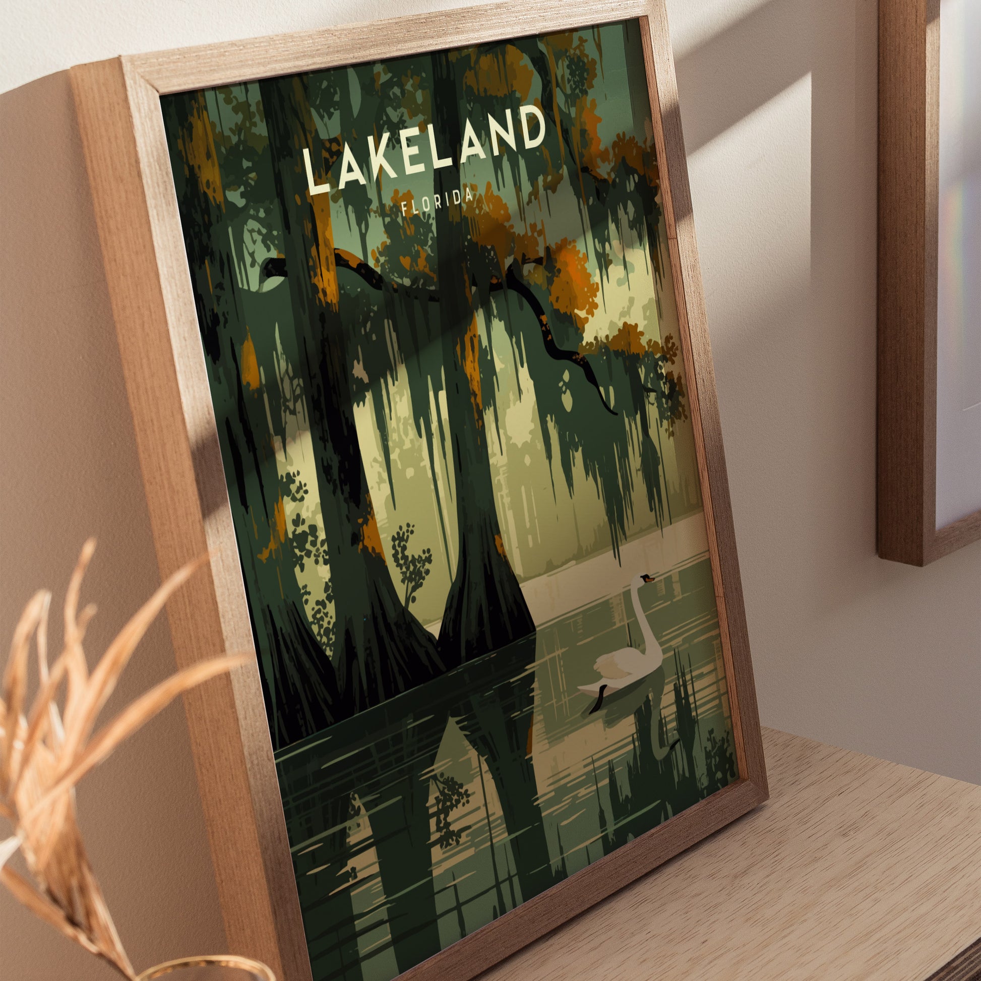 Framed poster featuring a serene illustration of Lake Hollingsworth with cypress trees and a swan, labeled Lakeland Florida, displayed on a wooden surface.