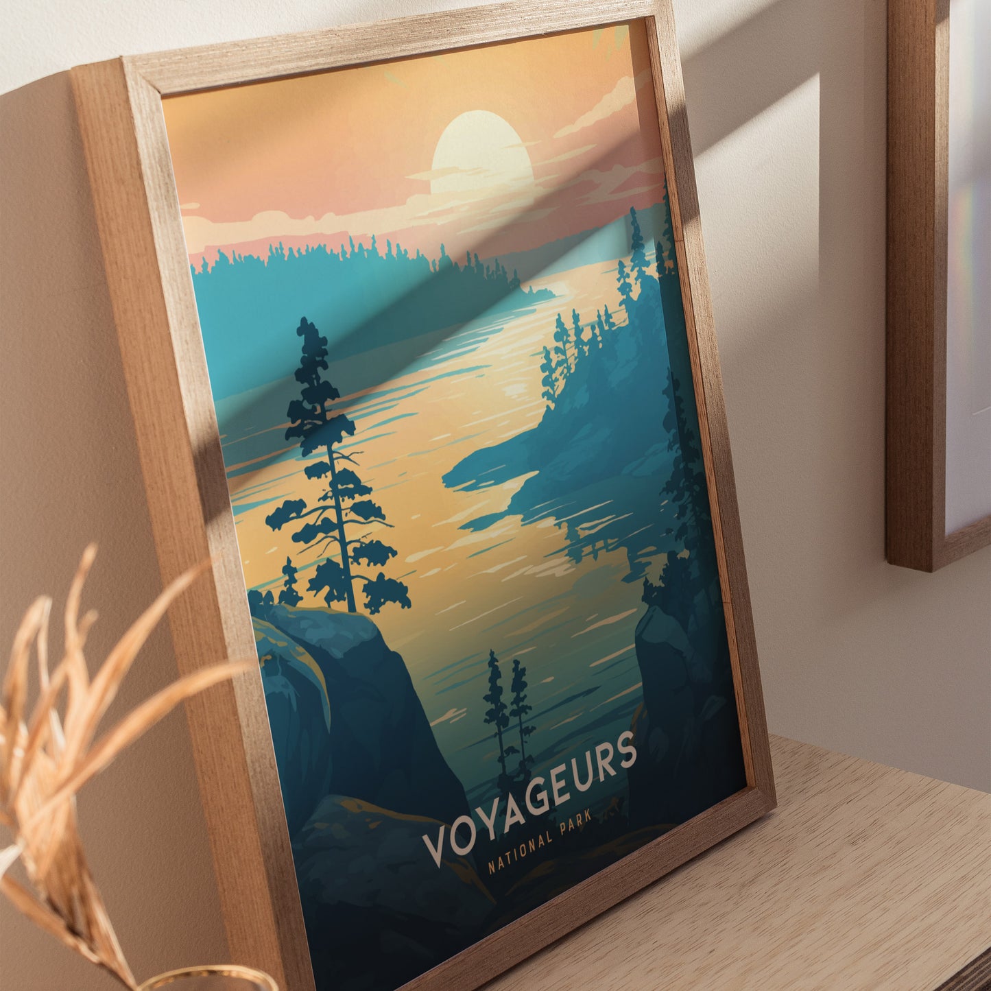 Framed poster depicting a serene lake scene at sunset with silhouetted trees and a glowing sky, labeled Voyageurs National Park. The artwork leans against a light-colored wall.