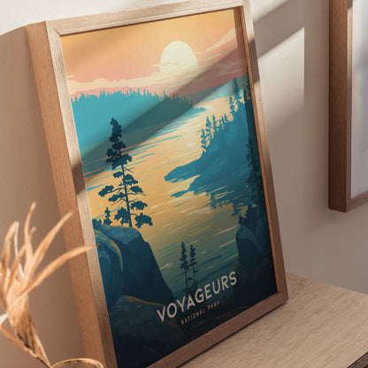 Framed poster depicting a serene lake scene at sunset with silhouetted trees and a glowing sky, labeled Voyageurs National Park. The artwork leans against a light-colored wall.