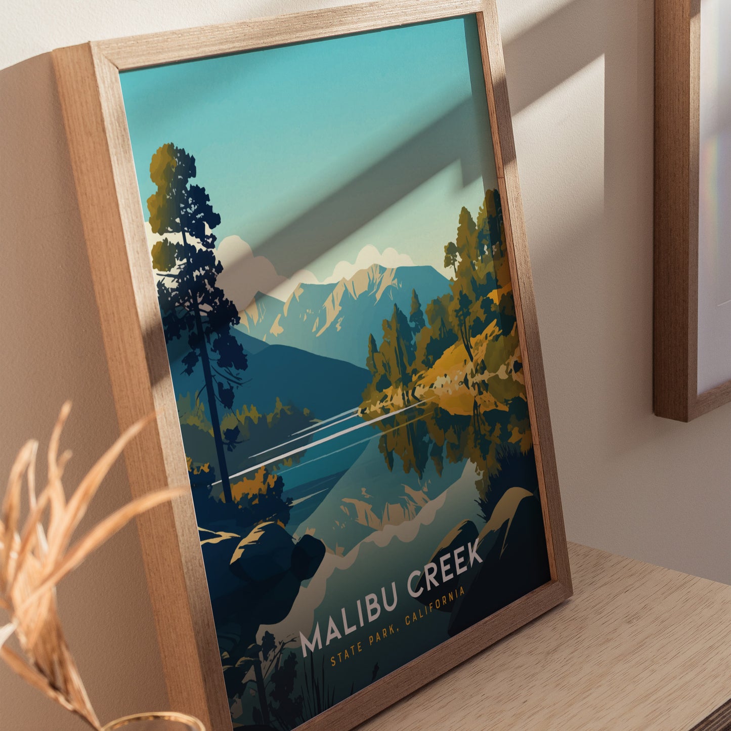 Framed poster depicting Malibu Creek State Park with stylized mountains, trees, and a winding creek, set on a wooden shelf. Natural light casts shadows on the artwork.