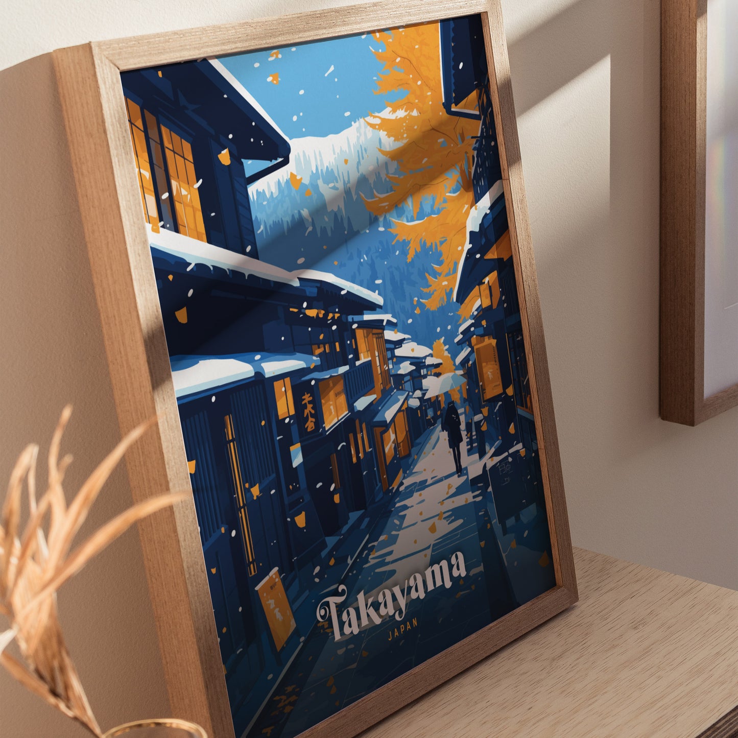 A framed poster of Takayama, Japan, featuring snow-covered traditional Edo-era streets with vibrant autumn leaves and distant mountains, leaning against a wall on a wooden surface.