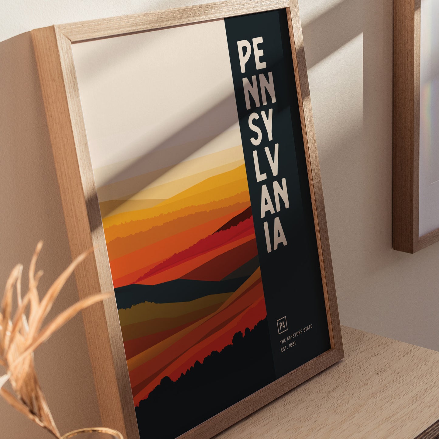 Framed Pennsylvania poster featuring abstract landscape in warm hues and vertical text. Placed on a wooden surface with soft lighting creating shadows.