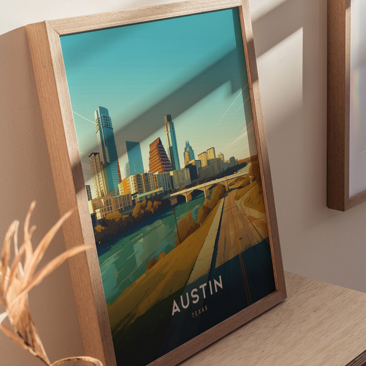 Framed artwork depicting the Austin, Texas skyline with modern buildings and a river under a clear blue sky, placed on a wooden surface with soft shadows.