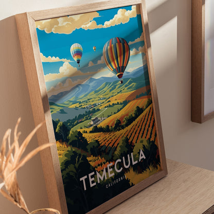 A framed poster of Temecula, California, depicting colorful hot air balloons over sunlit vineyards and rolling hills, is displayed on a light wooden surface.