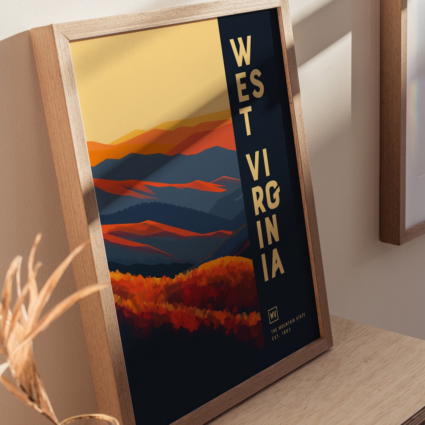 Framed poster of West Virginia featuring abstract, layered mountains in warm oranges and cool blues, with bold text on the side. Displayed on a wooden surface with natural lighting.