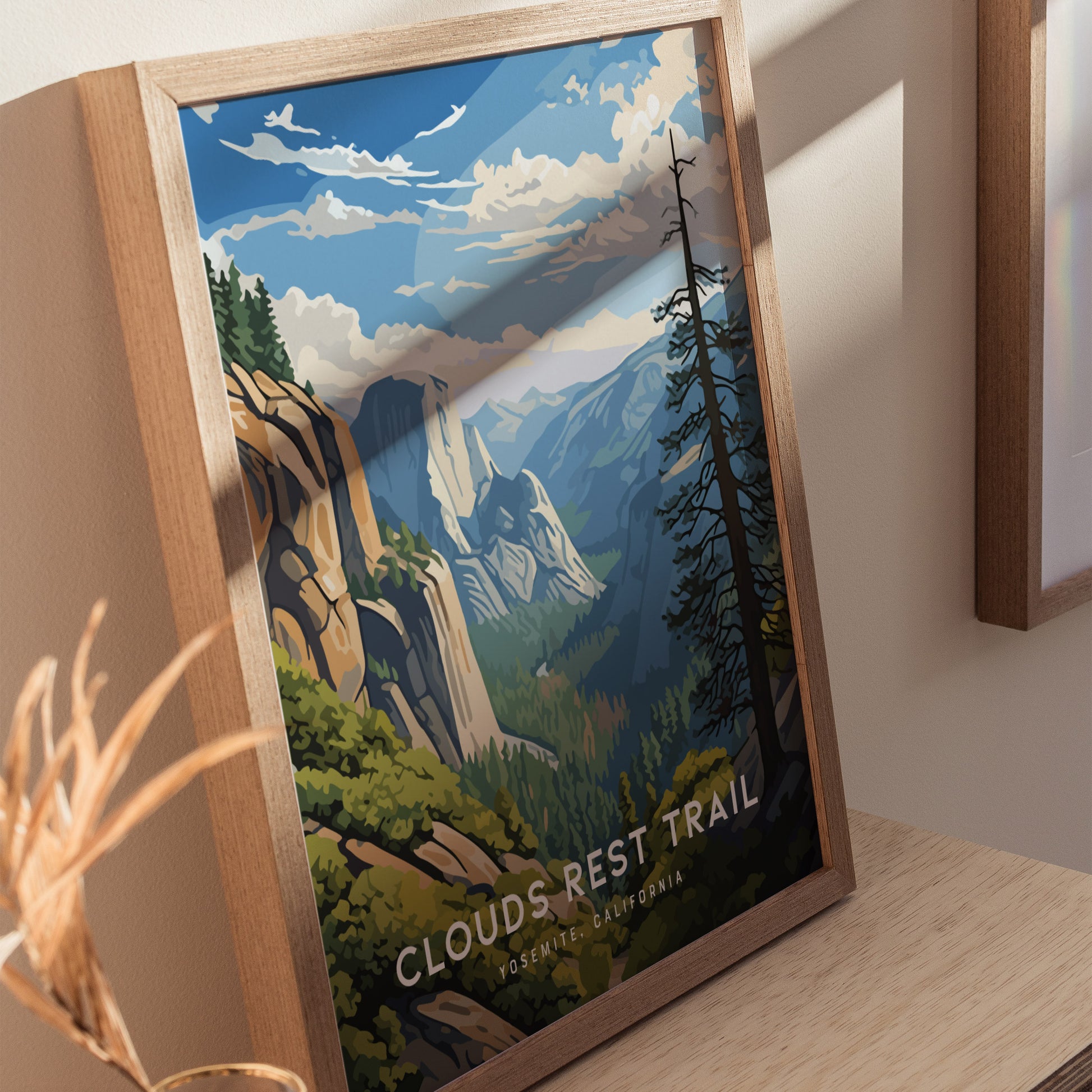 Framed artwork depicting a vibrant landscape of Yosemites Clouds Rest Trail, featuring towering granite cliffs, lush greenery, and a dramatic sky. The poster rests on a wooden surface.