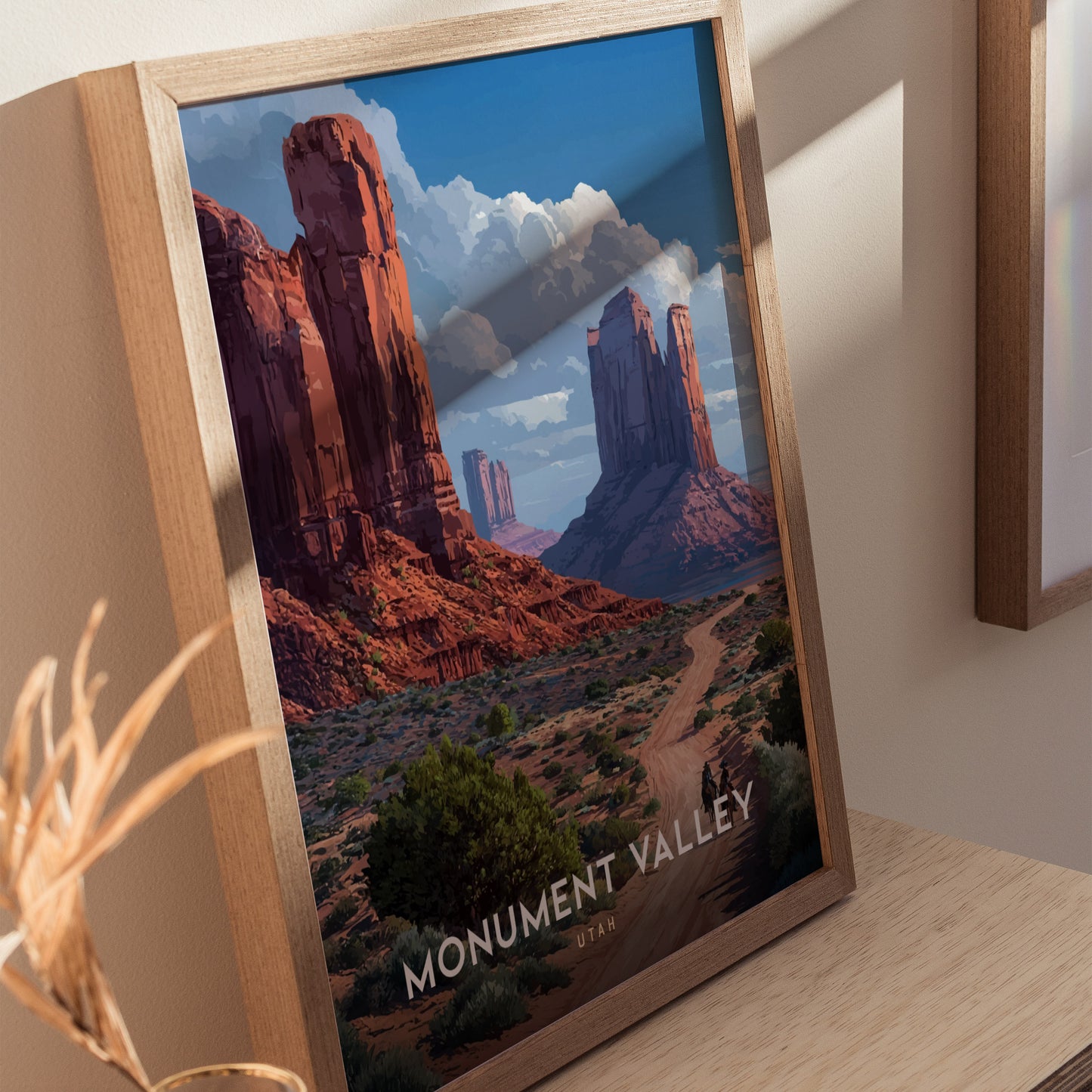 A framed poster of Monument Valley features red sandstone mesas and a winding desert path under a blue sky. The artwork is positioned on a wooden surface.