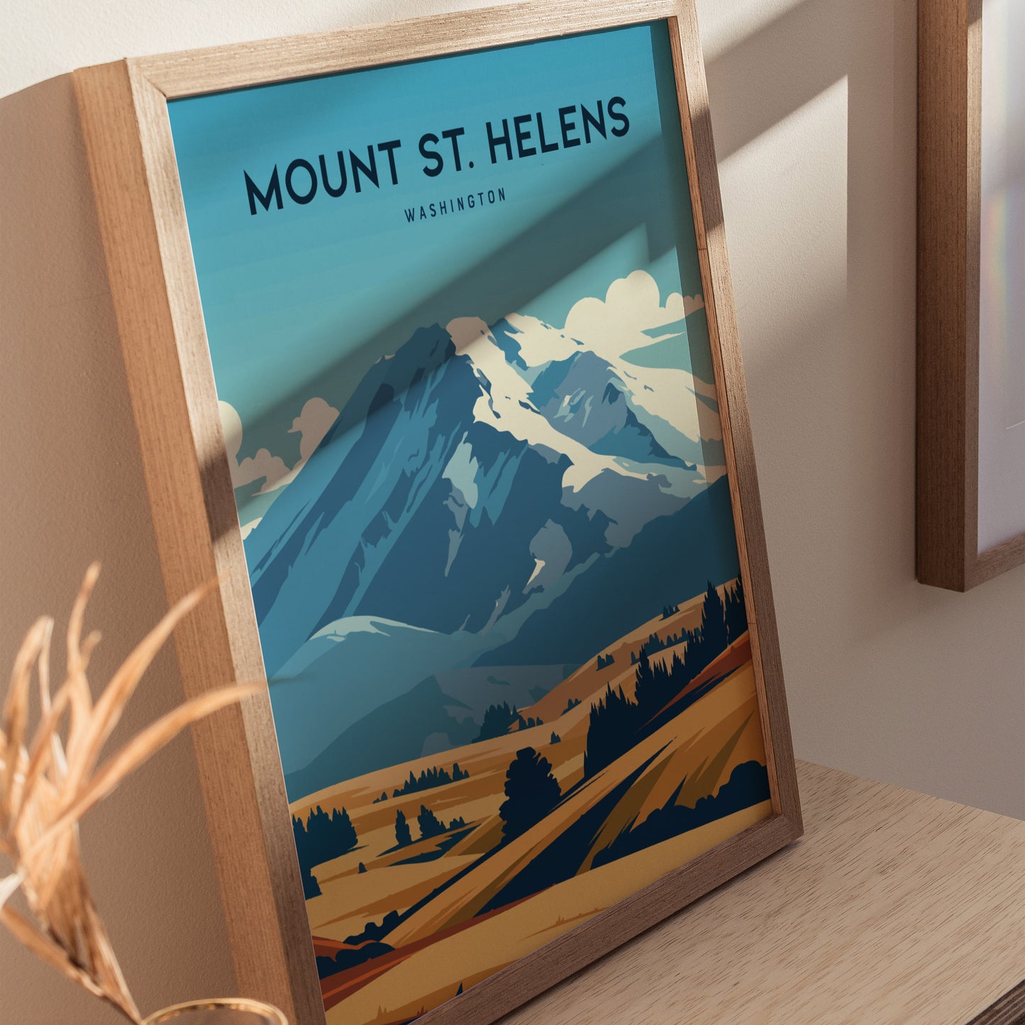 Framed Mount St. Helens poster depicting a stylized landscape with blue mountains and golden fields, placed on a wooden surface with soft lighting and shadows.