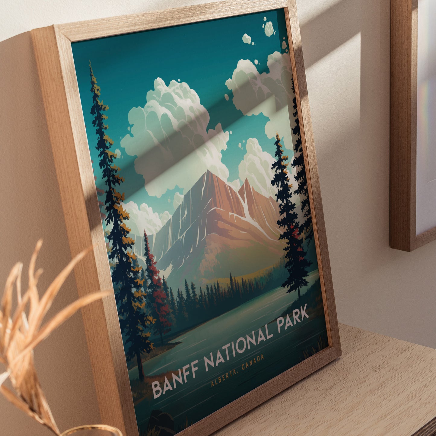 A framed poster of Banff National Park features a stylized mountain landscape with vibrant trees and clouds, set on a wooden surface with soft sunlight casting shadows.