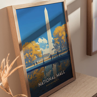 A framed poster of the National Mall in Washington D.C. features the Washington Monument, autumn trees, and a reflective water scene, displayed on a wooden surface.