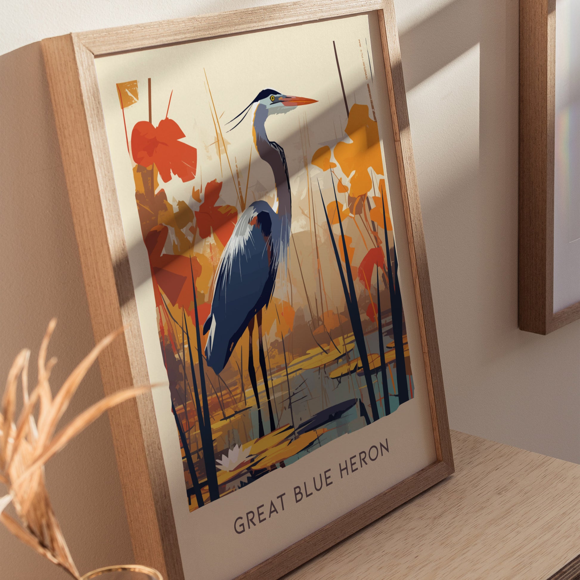 A framed poster of a Great Blue Heron stands on a wooden surface, featuring the bird among orange and yellow foliage. The sunlight casts a soft shadow on the wall.