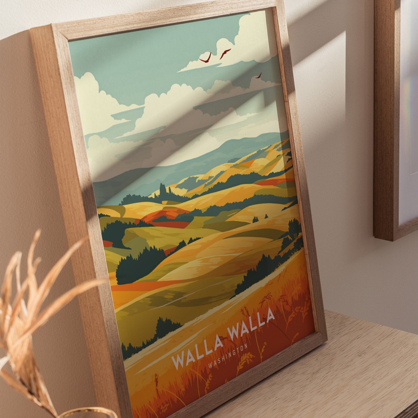 A framed poster featuring colorful, rolling hills under a partly cloudy sky with birds flying above, labeled Walla Walla Washington. The artwork is positioned on a wooden surface.