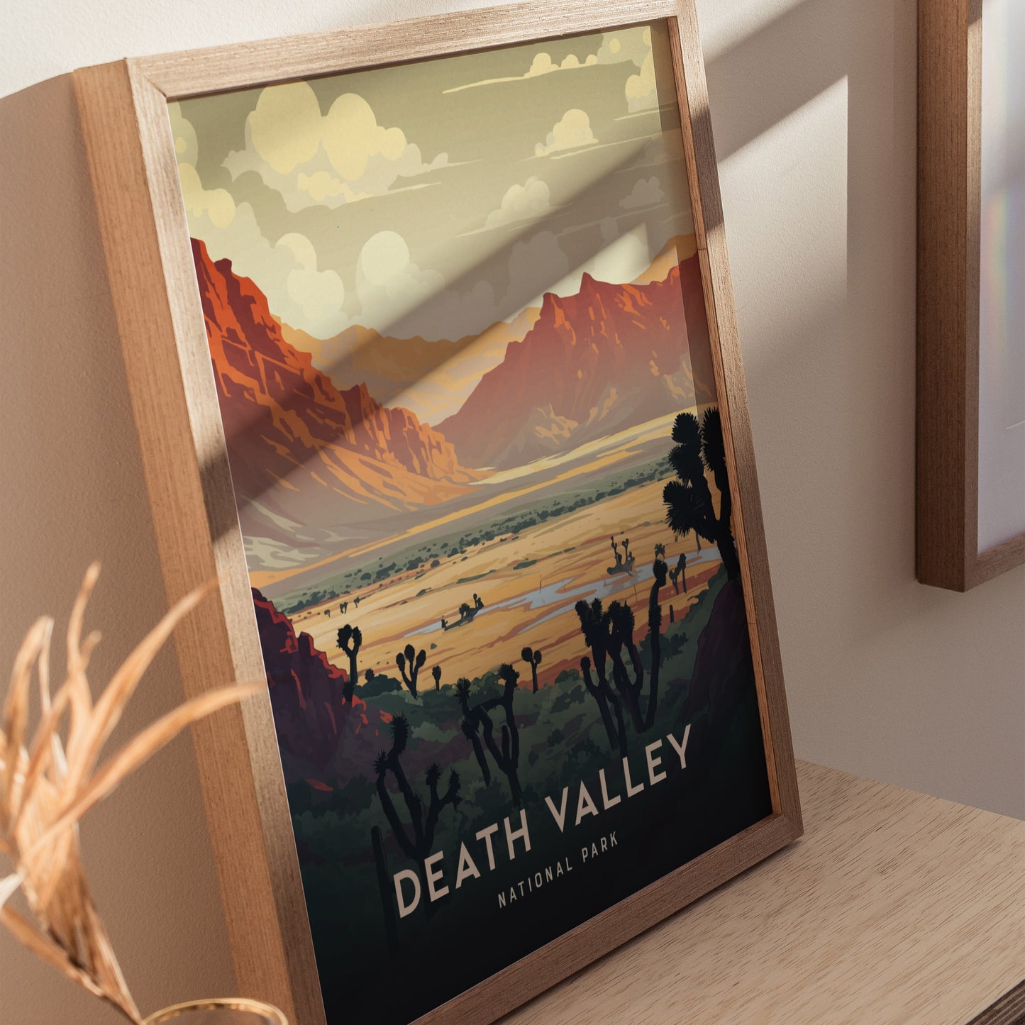 Framed art print of Death Valley National Park features a colorful landscape with rugged mountains, desert vegetation, and a dramatic sky. The wooden frame rests on a light wooden surface.