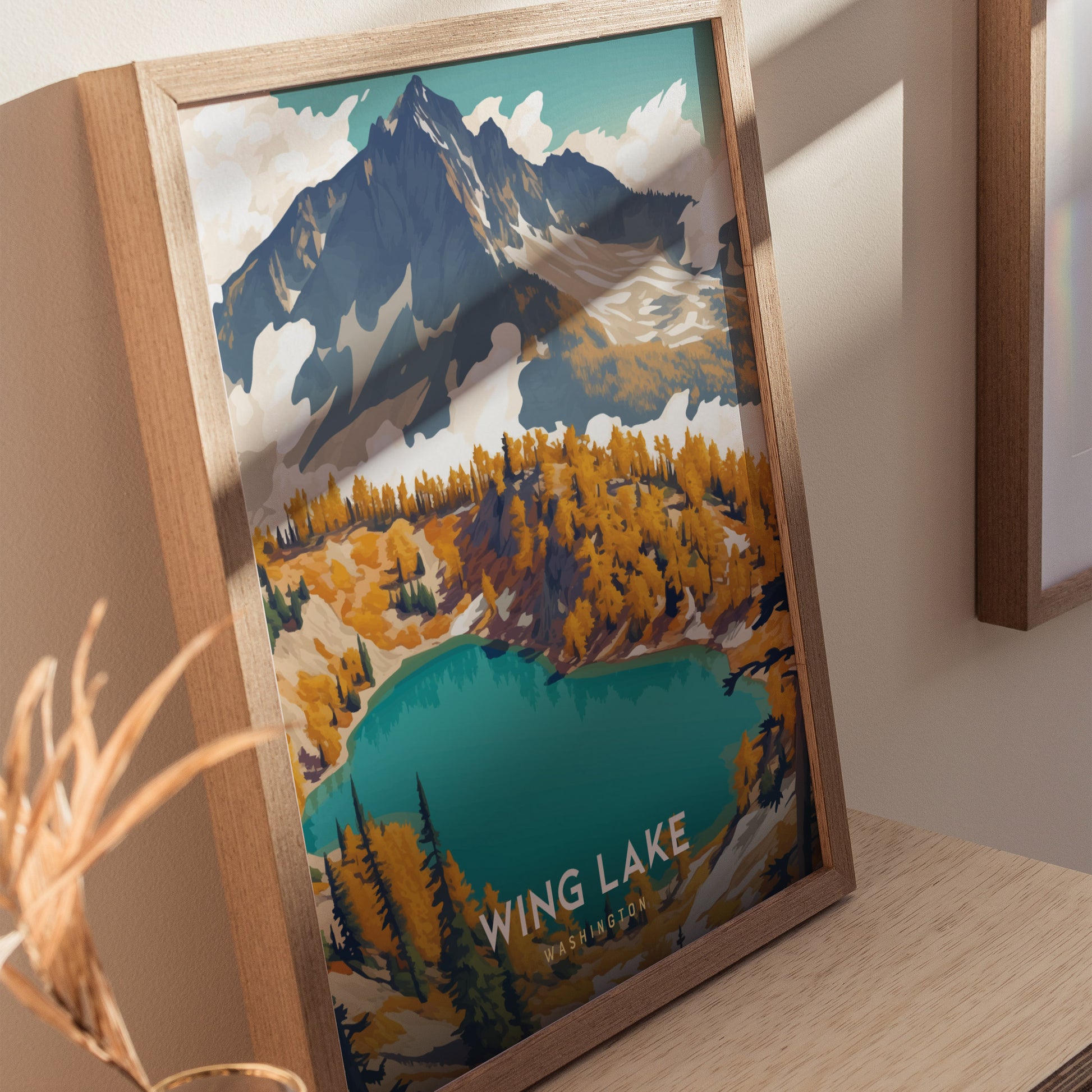 A framed poster of Wing Lake, Washington, showcasing vibrant turquoise waters surrounded by autumnal trees and a towering mountain, is displayed on a wooden surface.