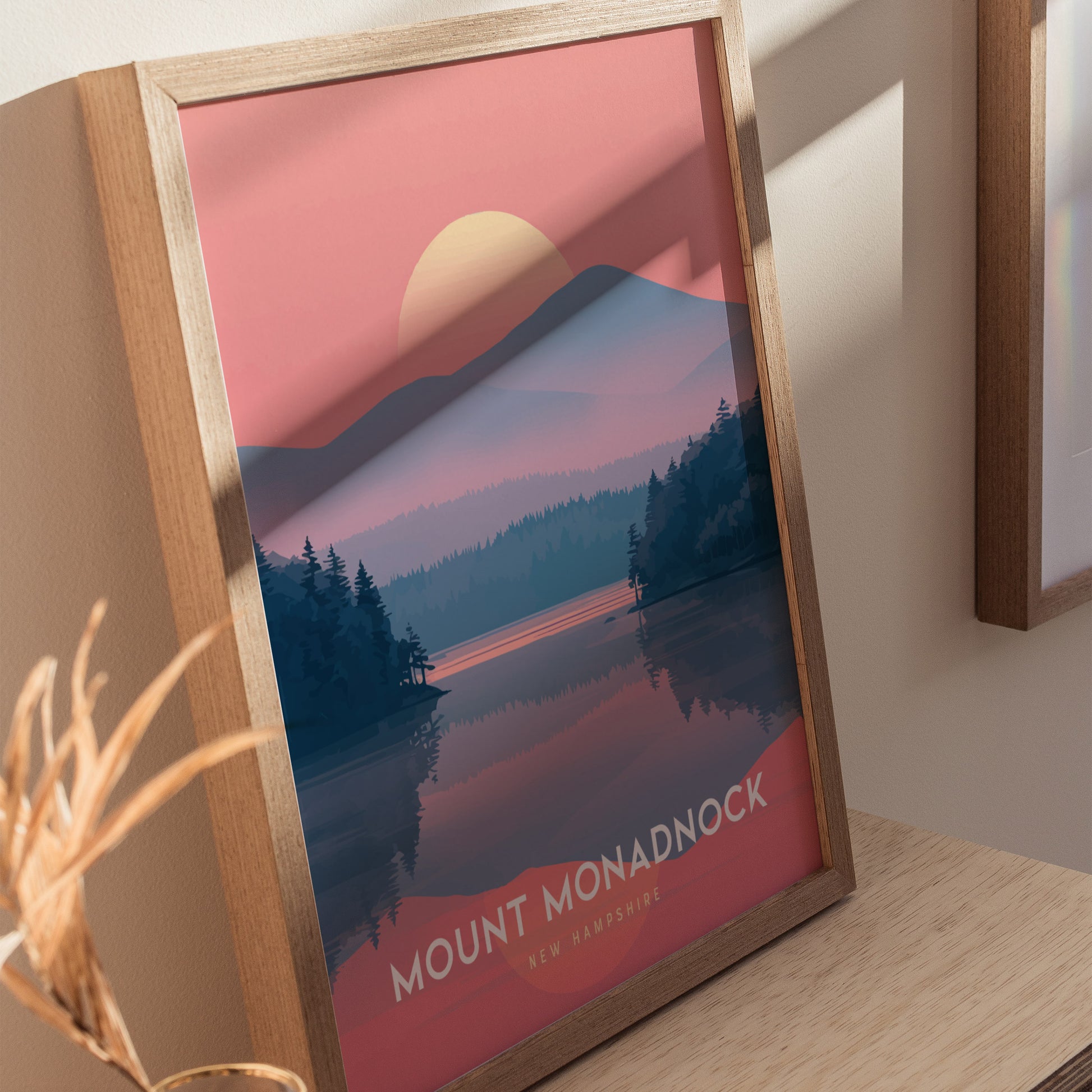 Framed Mount Monadnock poster with a sunset over mountains and a forest reflected in a lake. The artwork features a pink and blue color scheme and sits on a wooden.