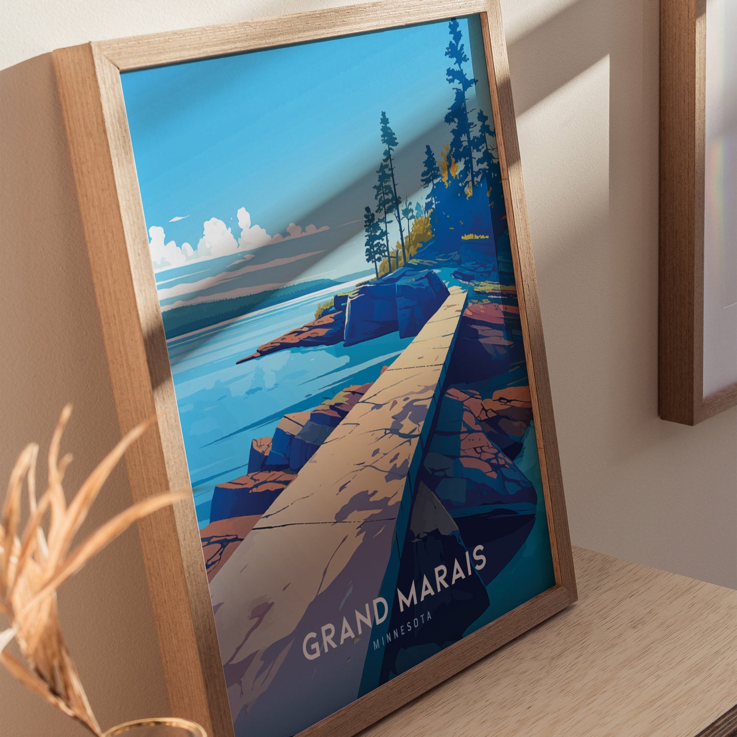 Framed artwork depicting a vibrant landscape of Grand Marais, Minnesota, featuring a rocky shoreline and pine trees under a clear blue sky. The poster rests against a light-colored wall.